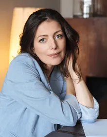 A picture of a dark haired woman woman sitting at a desk wearing a light blue blazer