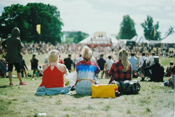 A crowd sits outside on the grass with their backs to the caera, looking towards a stage in the distance